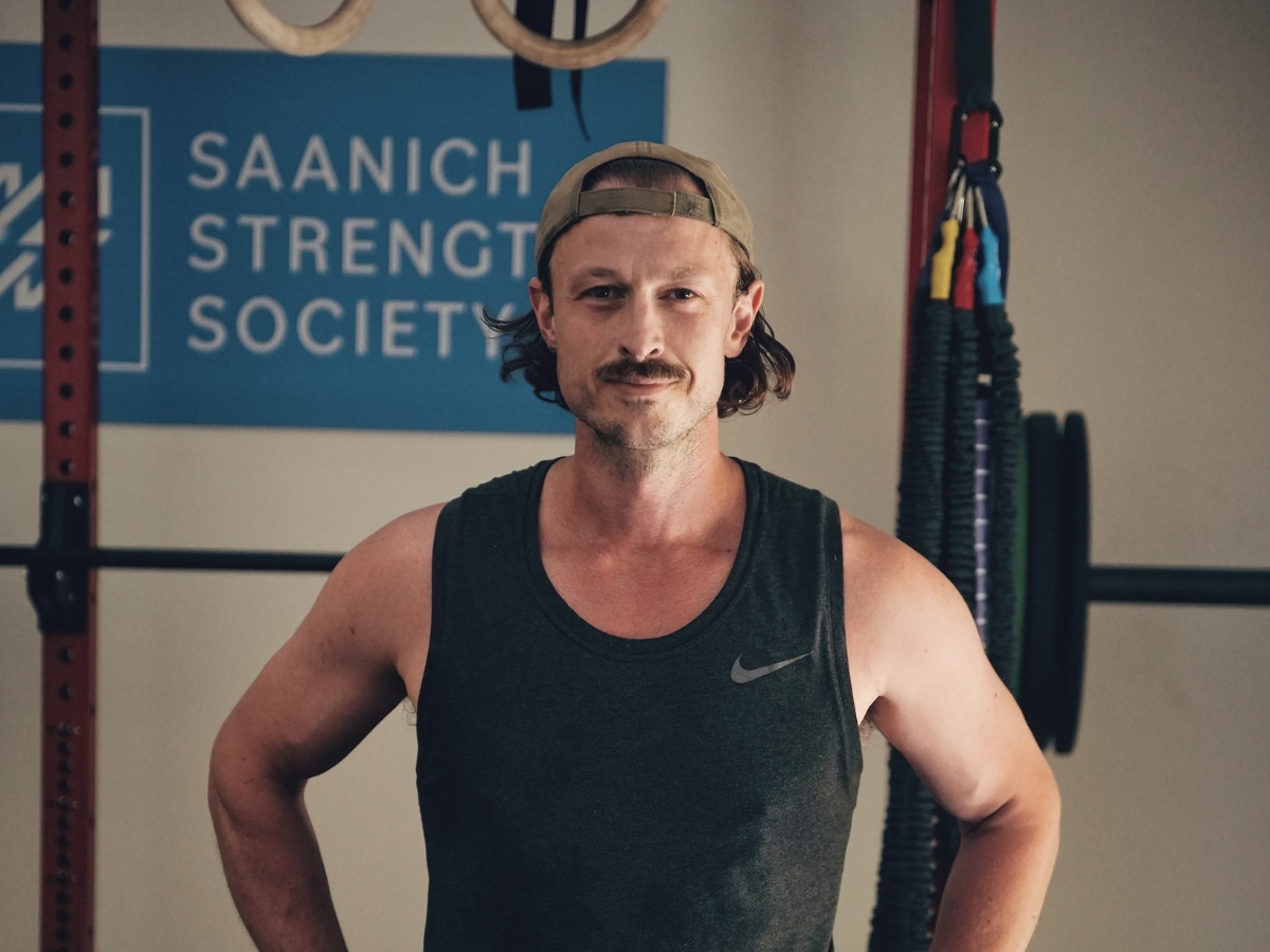 Alex standing in a gym with a black tank top on and a backwards hat with the sign reading Saanich Strength Society behind him.